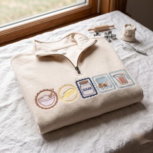 May include: A cream-colored quarter-zip sweatshirt featuring five embroidered baking-themed patches across the front. The designs include a mixing bowl, a stick of butter, a jar labeled SUGAR, a carton of eggs, and a measuring cup with clear markings.