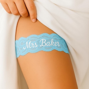 May include: A light blue lace garter with the text "Mrs Baker" in white script. The garter is worn on a leg, with a white dress visible in the background. The garter has a scalloped edge.