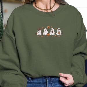 K&ouml;nnte beinhalten: Olivgr&uuml;nes Sweatshirt mit einem gestickten Design von vier Cartoon-Geistern. Jeder Geist hat ein einzigartiges Halloween-Accessoire, darunter ein Hexenhut, ein K&uuml;rbis und eine S&uuml;&szlig;igkeitent&uuml;te. Das Design umfasst Herbstbl&auml;tter und einen K&uuml;rbis.