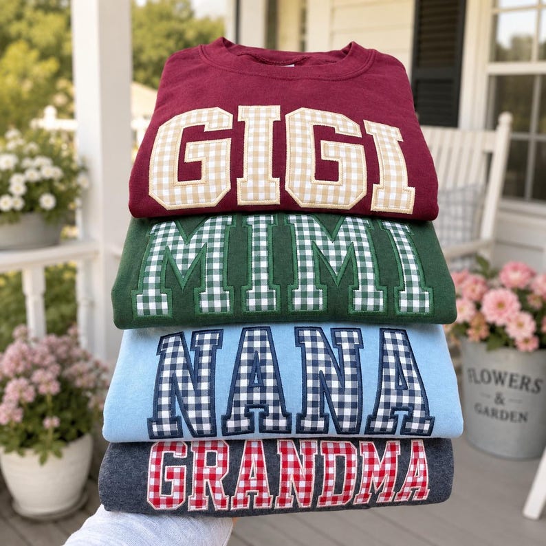 May include: A stack of four crewneck sweatshirts in maroon, dark green, light blue, and charcoal gray. Each sweatshirt features large, block-style letters appliqued in a gingham pattern, spelling out the words GIGI, MIMI, NANA, and GRANDMA.