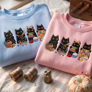 Op de afbeelding: Een lichtblauw en een lichtroze sweater met ronde hals, elk voorzien van een borduursel van vier zwarte katten die bezig zijn met naaien en knutselen, waaronder garen, scharen, stof en een quilt, met diverse naaibenodigdheden in de buurt.