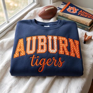 May include: A navy blue crewneck sweatshirt featuring the word AUBURN in large, block-style letters with varied orange and white patterns, including polka dots, stripes, gingham, and floral prints, with the word tigers in orange script below.