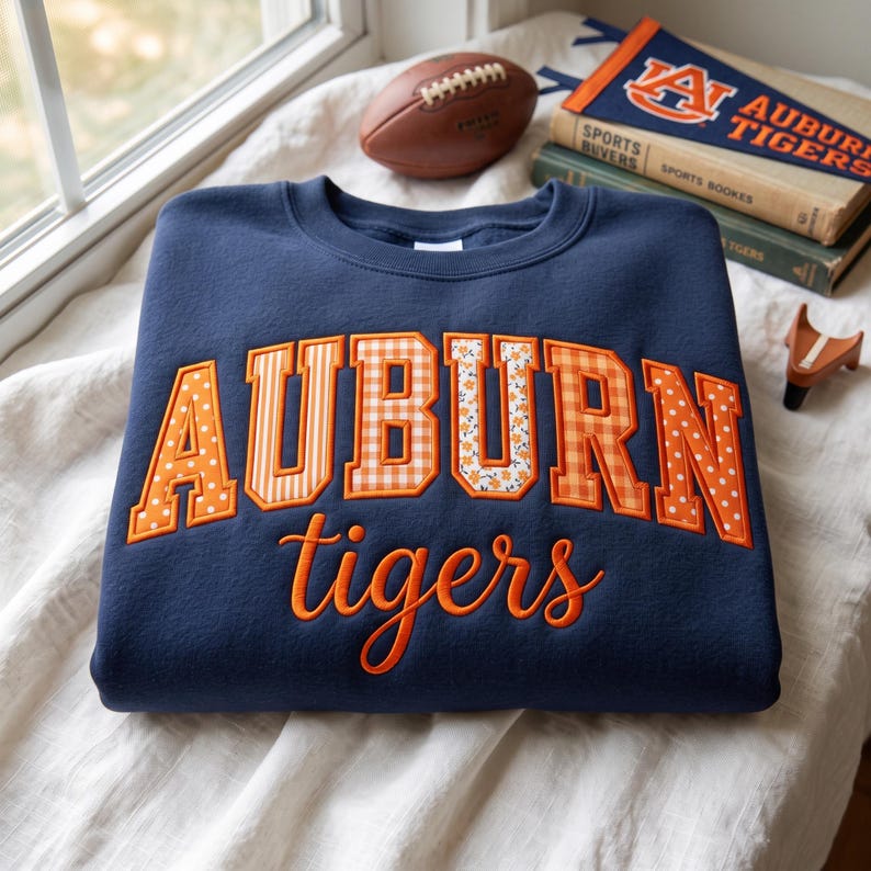 May include: A navy blue crewneck sweatshirt featuring the word AUBURN in large, block-style letters with varied orange and white patterns, including polka dots, stripes, gingham, and floral prints, with the word tigers in orange script below.