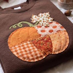 May include: A dark brown sweatshirt featuring a large, central pumpkin appliqué. The pumpkin is constructed from various fabric patches in shades of orange, including gingham, floral, and polka dot patterns, with a green embroidered vine and leaf.