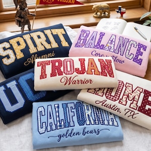 Könnte beinhalten: Eine Sammlung gefalteter Sweatshirts in verschiedenen Farben wie Marineblau, Rosa, Weiß und Hellblau. Jedes Sweatshirt zeigt große Blockbuchstaben mit gemusterten Stofffüllungen und darunter kleineren Schreibschrifttext, wie SPIRIT Alumni, BALANCE Core Flow, TROJAN Warrior, HOME Austin, TX und CALIFORNIA golden bears.