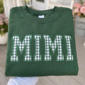 May include: A dark forest green crewneck sweatshirt featuring the word MIMI appliqued across the chest in a white and green gingham check pattern. The letters are block-style with a stitched border, centered on the front of the garment.
