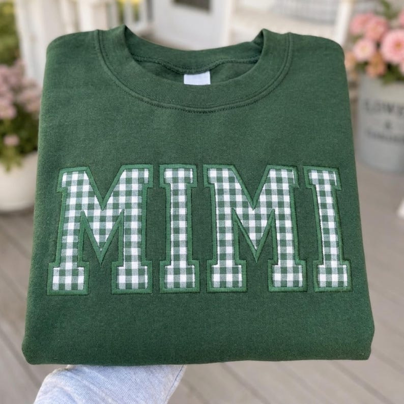 May include: A dark forest green crewneck sweatshirt featuring the word MIMI appliqued across the chest in a white and green gingham check pattern. The letters are block-style with a stitched border, centered on the front of the garment.
