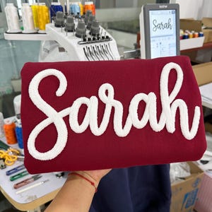 May include: A deep red sweatshirt featuring the name Sarah embroidered in large, white, textured chenille-style script lettering across the front. The fabric has a smooth finish, and the white letters stand out with a raised, fuzzy appearance.