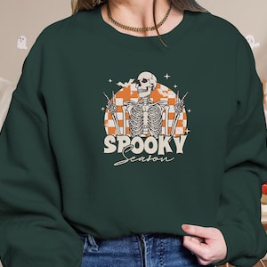 Pode incluir: Camisola verde escuro com um gráfico retro. O gráfico apresenta um esqueleto a fazer o sinal de paz, contra um fundo xadrez laranja e branco. As palavras "SPOOKY Season" estão escritas abaixo do esqueleto numa fonte vintage.