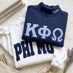 Peut inclure: Un sweat-shirt bleu marine à col montant avec des lettres grecques bleu clair à pois et le nom Sophia brodé sur le col, superposé sur un sweat-shirt blanc avec des lettres bleu foncé à pois épelant PHI MU dans une police universitaire.