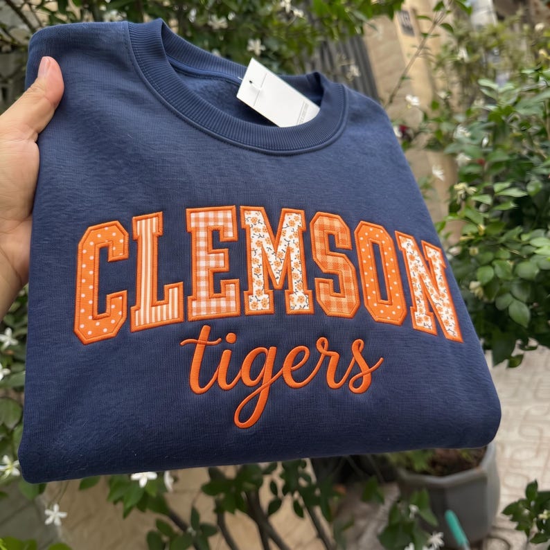 May include: A navy blue crewneck sweatshirt featuring the word CLEMSON in large, orange-bordered block letters with varied patterns including polka dots, stripes, and florals. Below, the word tigers is embroidered in orange cursive script.
