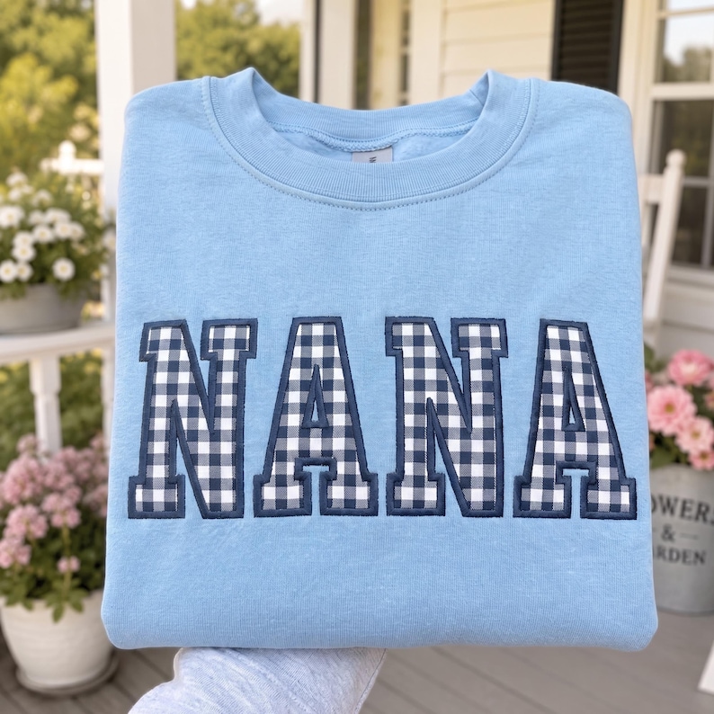 May include: A light blue crewneck sweatshirt featuring the word NANA appliqued across the chest in a navy blue and white gingham check pattern. The letters are outlined in a dark navy blue thread, creating a bold, block-style font design.