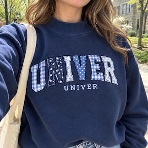 Puede incluir: Una sudadera azul marino de cuello redondo con la palabra UNIVER en letras grandes aplicadas. Cada letra tiene un patrón único en azul y blanco, incluyendo cuadros vichy, estrellas, lunares y flores. La palabra UNIVER está bordada debajo.