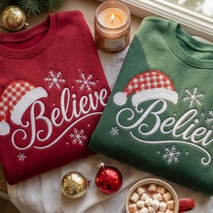 May include: A red sweatshirt and a green sweatshirt, each featuring white embroidered text that reads Believe, a red and white checkered Santa hat graphic, and white snowflake accents. The sweatshirts are displayed with holiday ornaments.