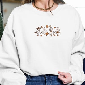 K&ouml;nnte beinhalten: Wei&szlig;es Sweatshirt mit einem gestickten Design von vier Cartoon-Geistern mit Halloween-Accessoires. Ein Geist tr&auml;gt einen Hexenhut, ein anderer h&auml;lt einen K&uuml;rbisballon und die anderen halten K&uuml;rbisse. Braune Bl&auml;tter sind verstreut.