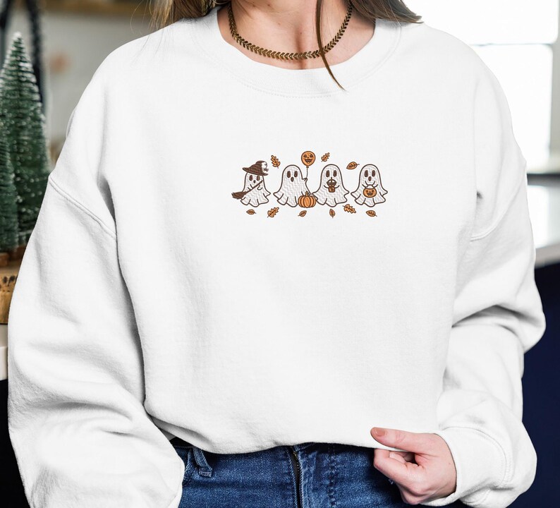 K&ouml;nnte beinhalten: Wei&szlig;es Sweatshirt mit einem gestickten Design von vier Cartoon-Geistern mit Halloween-Accessoires. Ein Geist tr&auml;gt einen Hexenhut, ein anderer h&auml;lt einen K&uuml;rbisballon und die anderen halten K&uuml;rbisse. Braune Bl&auml;tter sind verstreut.