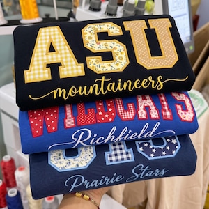 Könnte beinhalten: Ein Stapel aus drei Sweatshirts. Das oberste ist schwarz mit gelbem ASU-Schriftzug und dem Wort mountaineers. Das mittlere ist blau mit rotem WILDCATS-Schriftzug und dem Wort Richfield. Das unterste ist marineblau mit blauem Prairie Stars-Schriftzug.