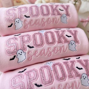 Op de afbeelding: Een lichtroze sweatshirt met de geborduurde tekst SPOOKY in grote glitterende roze blokletters en season in cursief schrift. Het ontwerp bevat kleine witte spookfiguren en zwarte vleermuissilhouetten verspreid over de stof.