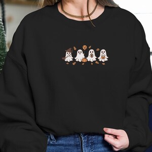 K&ouml;nnte beinhalten: Schwarzes Sweatshirt mit einem gestickten Design von vier Cartoon-Geistern. Die wei&szlig;en Geister haben gestickte Details und halten jeweils einen Halloween-Gegenstand. Herbstbl&auml;tter und ein Hexenhut vervollst&auml;ndigen das festliche Design.