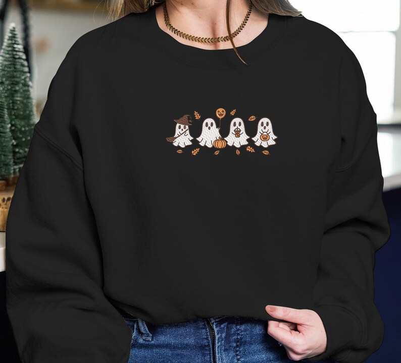 K&ouml;nnte beinhalten: Schwarzes Sweatshirt mit einem gestickten Design von vier Cartoon-Geistern. Die wei&szlig;en Geister haben gestickte Details und halten jeweils einen Halloween-Gegenstand. Herbstbl&auml;tter und ein Hexenhut vervollst&auml;ndigen das festliche Design.