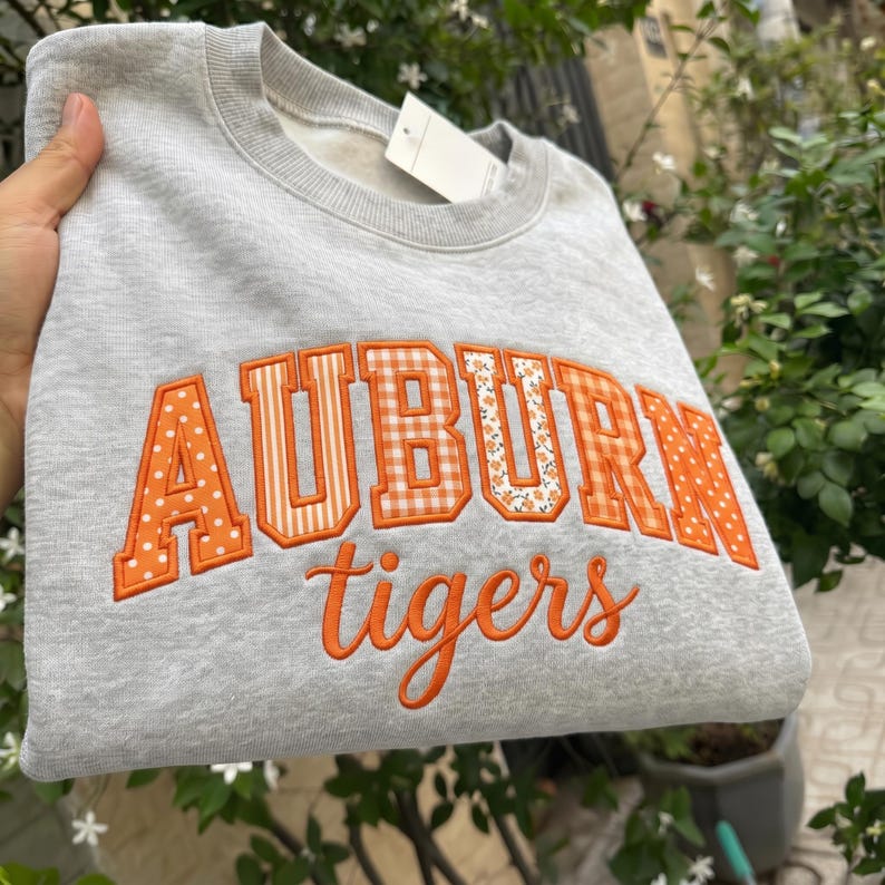 May include: A light gray crewneck sweatshirt featuring the word AUBURN in large, orange, patterned block letters with varying designs including polka dots, stripes, and florals. Below, the word tigers is embroidered in orange cursive script.
