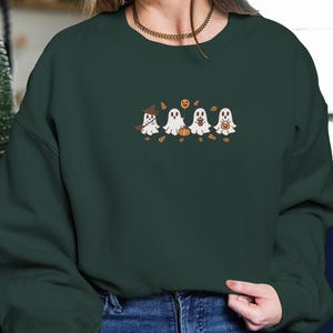 K&ouml;nnte beinhalten: Dunkelgr&uuml;nes Sweatshirt mit einem gestickten Design von vier Cartoon-Geistern. Ein Geist tr&auml;gt einen Hexenhut, die anderen halten K&uuml;rbisse oder S&uuml;&szlig;igkeitent&uuml;ten. Das Design umfasst Herbstbl&auml;tter und einen K&uuml;rbis.