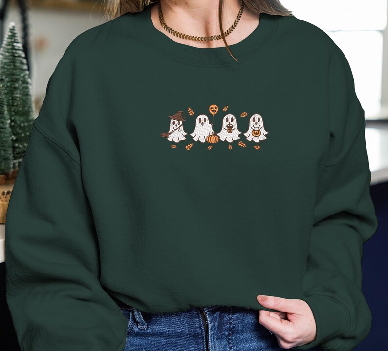 K&ouml;nnte beinhalten: Dunkelgr&uuml;nes Sweatshirt mit einem gestickten Design von vier Cartoon-Geistern. Ein Geist tr&auml;gt einen Hexenhut, die anderen halten K&uuml;rbisse oder S&uuml;&szlig;igkeitent&uuml;ten. Das Design umfasst Herbstbl&auml;tter und einen K&uuml;rbis.