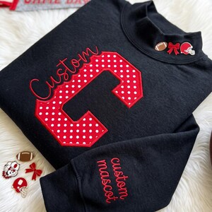 Puede incluir: Una sudadera negra de manga larga con una letra C roja grande con lunares blancos y la palabra Custom bordada en letra roja. El cuello muestra un balón de fútbol, un lazo rojo y un casco, mientras que la manga tiene el texto custom mascot.
