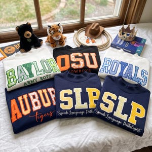 Peut inclure: Une collection de six sweat-shirts à col rond en blanc, noir et bleu marine, chacun arborant de grandes lettres en bloc avec des motifs variés. Les designs incluent des textes tels que BAYLOR, OSU, ROYALS, AUBURN et SLP Speech Language Pathologist.