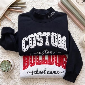 Könnte beinhalten: Ein schwarzes Sweatshirt mit Stehkragen, weißer gepunkteter CUSTOM-Schrift und dem Namen Sophia am Kragen. Darunter liegt ein hellgraues Sweatshirt mit roter gepunkteter BULLDOG-Schrift und dem Text school name in Schreibschrift.