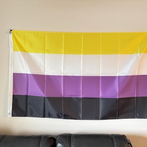 May include: A non-binary pride flag, rectangular in shape, hangs on a wall. The flag features horizontal stripes of yellow, white, purple, and black. The flag is made of a lightweight material and is mounted with grommets.