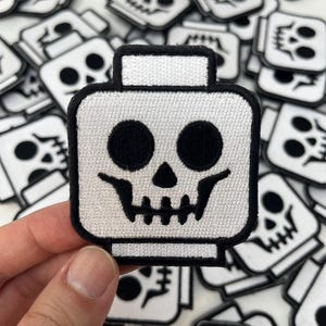 May include: White embroidered patch of a skull with black eyes and a black outline. The skull is shaped like a Lego head.