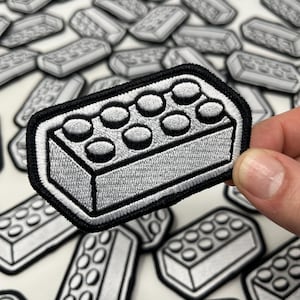 May include: Embroidered patch in the shape of a grey building block with a black border. The patch features a detailed design with circular studs on top. Multiple patches are scattered on a white surface.