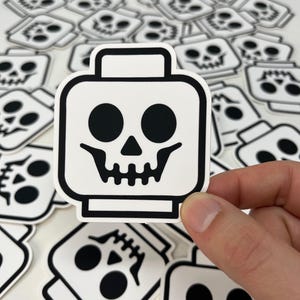 May include: A collection of white stickers with a black skull design. The skull is stylized with large eye sockets and a detailed jawline, set against a white background. The stickers are in the shape of a block head.