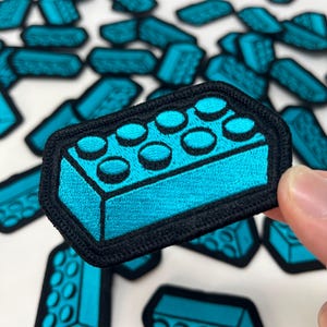 May include: Teal building block patches with a black border, scattered on a white surface. One patch is held by a hand. The patches are embroidered.