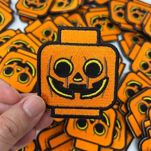May include: Orange embroidered patch in the shape of a jack-o'-lantern with black eyes and a smiling mouth. The patch has a black border and a rectangular top. Many similar patches are scattered in the background.