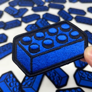 May include: A blue embroidered patch in the shape of a classic building block, with a black border. The patch features a detailed design with raised circular studs. Other similar patches are scattered in the background.