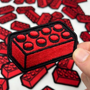 May include: Red embroidered patch in the shape of a classic building block, with a black border. The patch features a textured surface and circular studs. Several similar patches are scattered in the background.