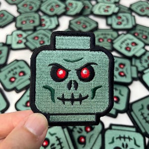 May include: A square-shaped embroidered patch featuring a light green stylized head with a black border. The design includes two glowing red eyes, a small nose, and a stitched mouth line, resembling a block-style toy character head.