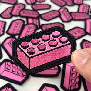 May include: Pink embroidered patches in the shape of a classic building brick. Each patch has a black border and features the brick's iconic circular studs. The patches are scattered across a white surface, with one patch being held.