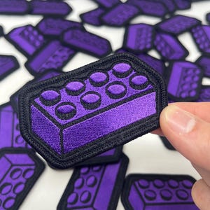 May include: Purple embroidered patch in the shape of a building block with black trim. The patch has a textured appearance and is held by a hand. Other patches are scattered in the background.