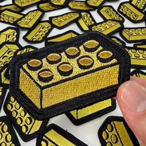 May include: A rectangular embroidered patch featuring a gold-colored building block design with eight raised circular studs on top, outlined by a thick black border. The patch is shown in a collection of identical pieces on a white surface.