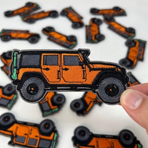May include: An orange and black embroidered patch depicting a Jeep-style vehicle. The patch has black tyres, a black roof, and a green accent. Several similar patches are scattered around the main patch.