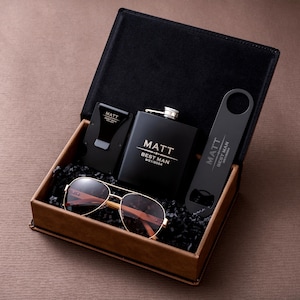 May include: A black gift box containing a hip flask, sunglasses, a money clip, and a bottle opener. The flask and bottle opener are black with the name "Matt" and the text "Best Man 9/21/2024". The sunglasses have gold frames and brown lenses.