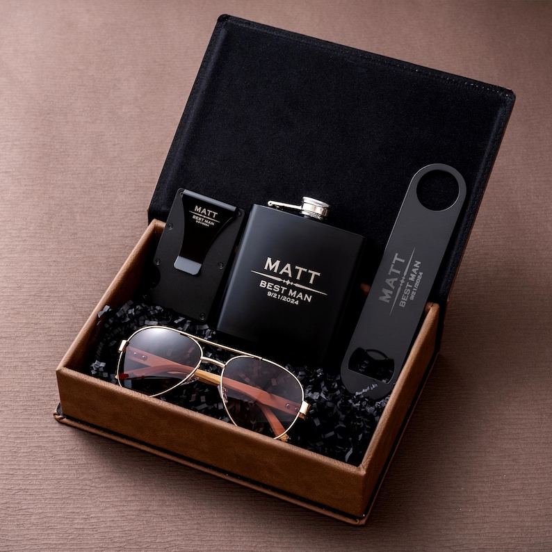 May include: A black gift box containing a hip flask, sunglasses, a money clip, and a bottle opener. The flask and bottle opener are black with the name "Matt" and the text "Best Man 9/21/2024". The sunglasses have gold frames and brown lenses.