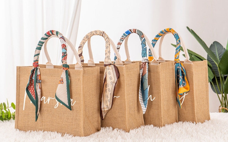May include: Five brown burlap tote bags with white handles and personalized names. Each bag has a colorful patterned scarf tied around the handles.