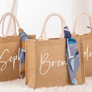 May include: Three brown burlap tote bags with white handles and personalized names. The bags are decorated with colorful patterned scarves.