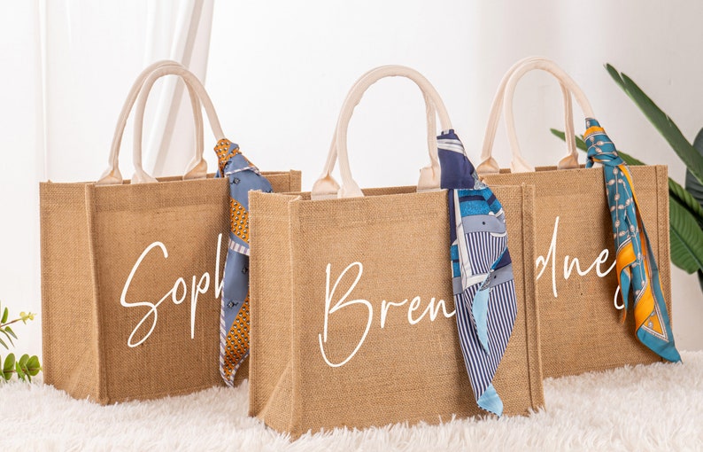 May include: Three brown burlap tote bags with white handles and personalized names. The bags are decorated with colorful patterned scarves.