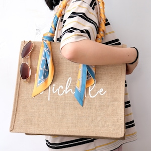 May include: A large, brown, burlap tote bag with a white inscription that reads "Michelle". The bag has a blue and yellow patterned scarf tied to the handles.