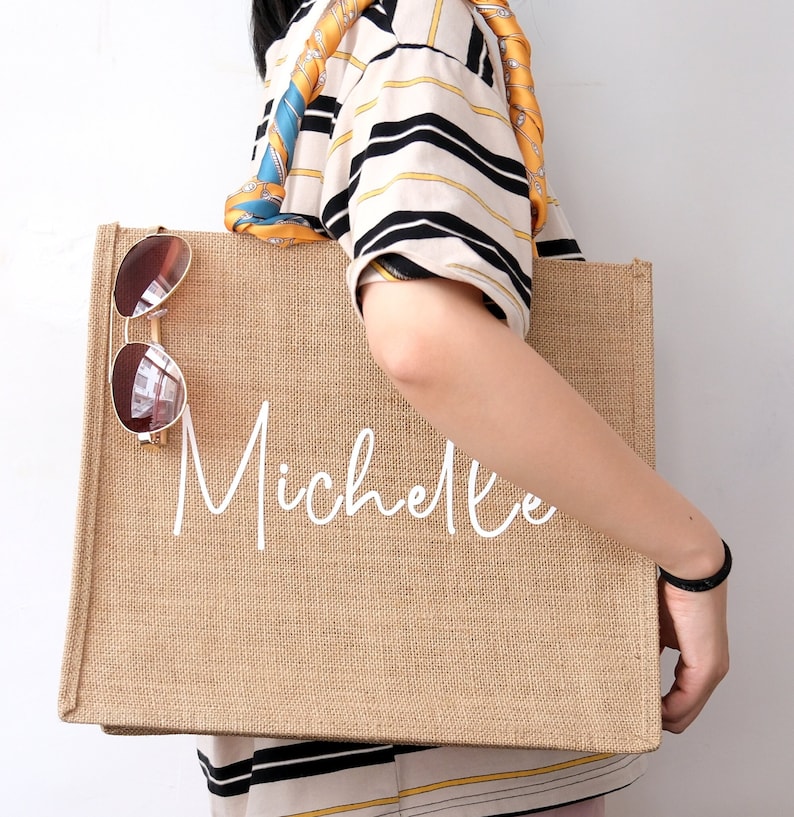 May include: A brown burlap tote bag with the name "Michelle" printed in white. The bag has a long strap and is being held by a person wearing a striped shirt.
